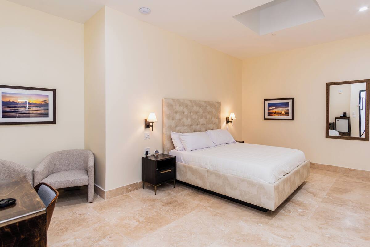 Un dormitorio con cama, mesitas de noche, una silla y dos cuadros en la pared.