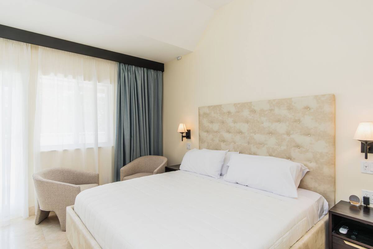 Habitación de hotel con una cama grande, una silla, una mesita de noche con lámpara y una luz en la pared.