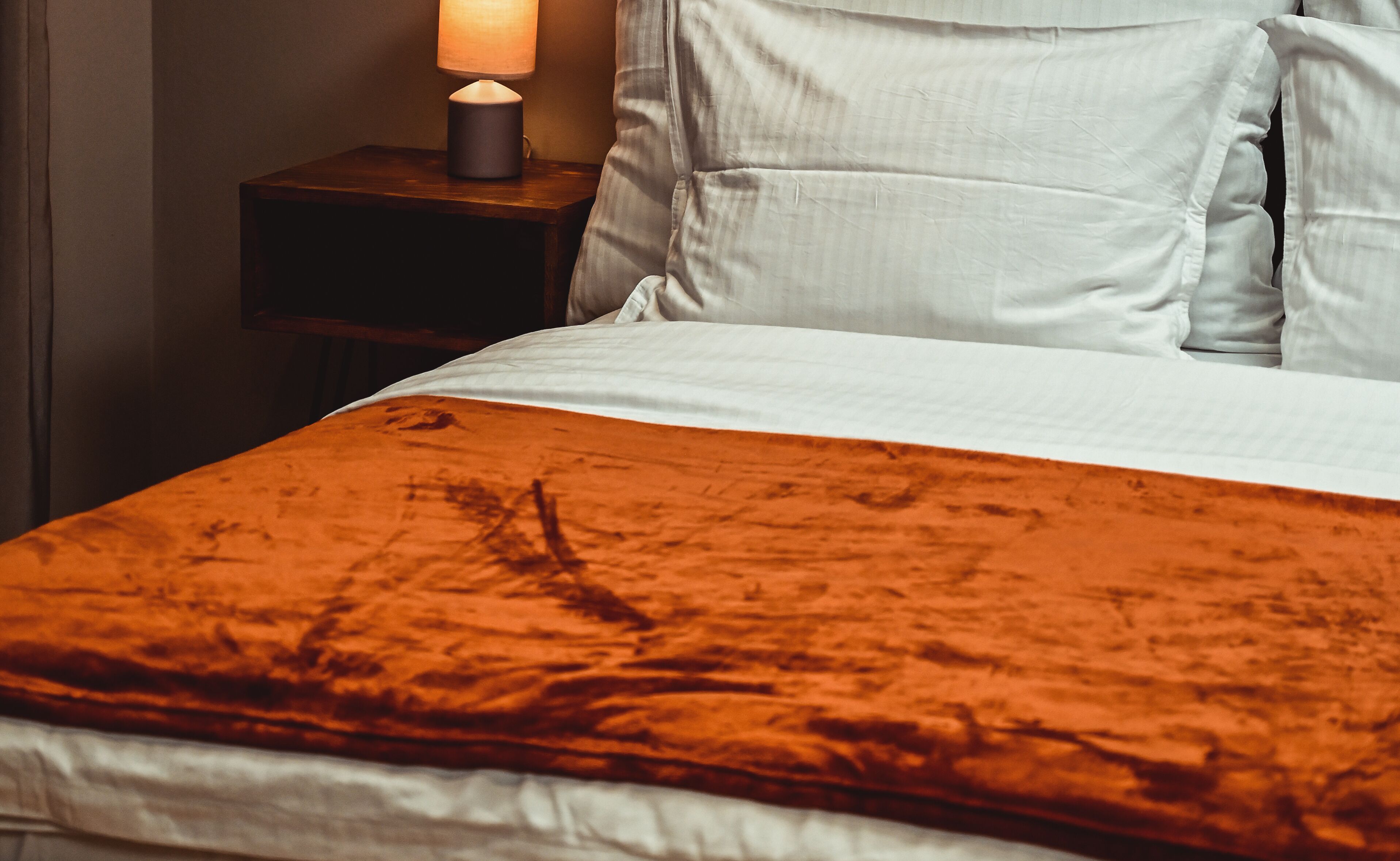 A bed with a white sheet and an orange blanket. There is a nightstand with a lamp beside the bed.