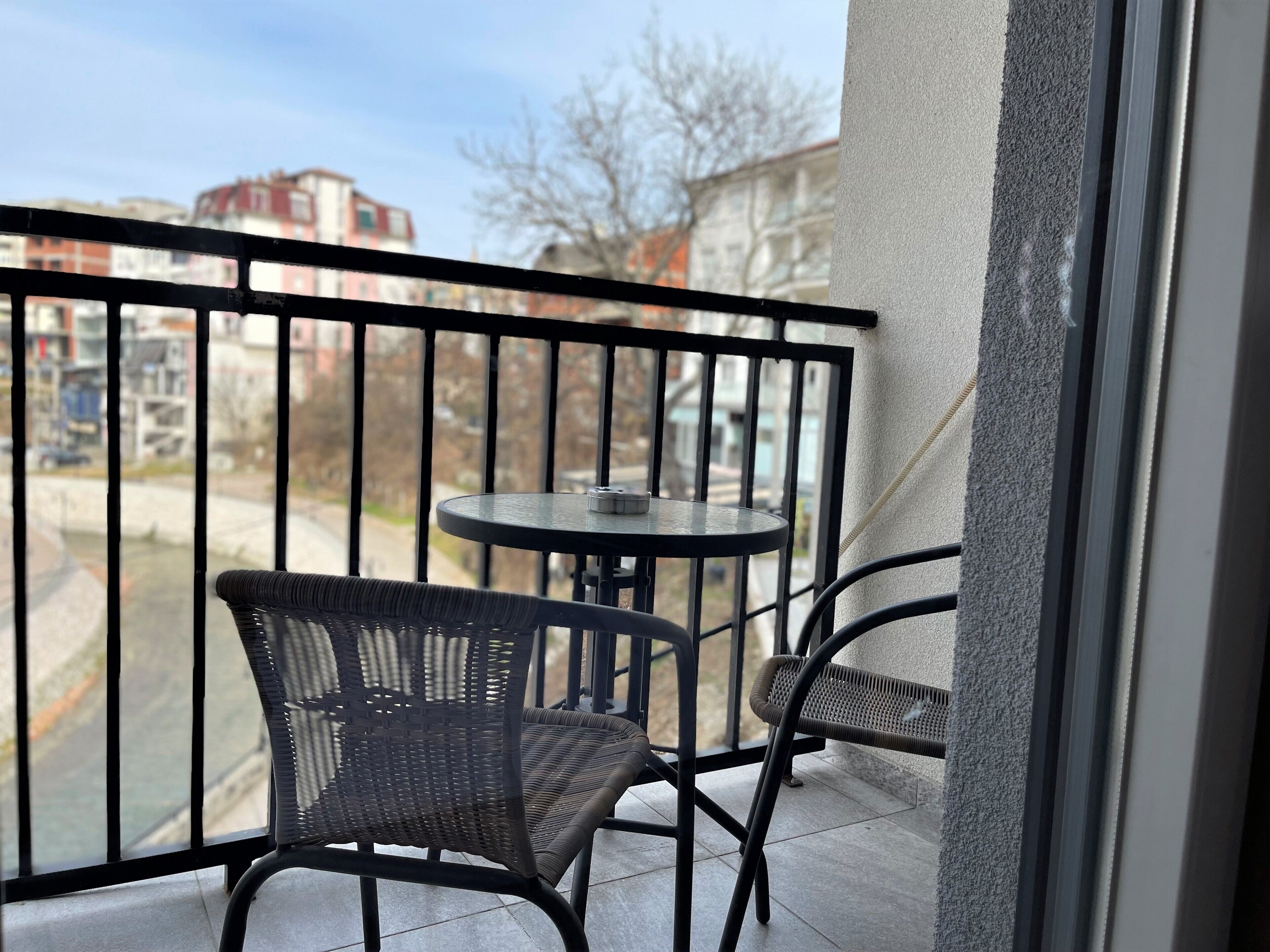 A balcony with two wicker chairs and a small round table, overlooking a street with buildings and trees.