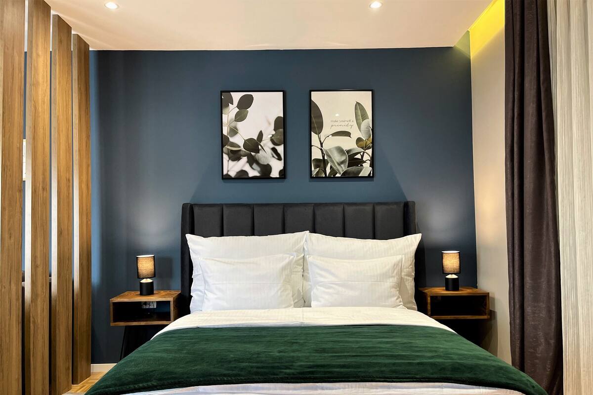 Une chambre avec un mur bleu foncé, une tête de lit noire, du linge de lit blanc et deux tables de chevet avec des lampes.
