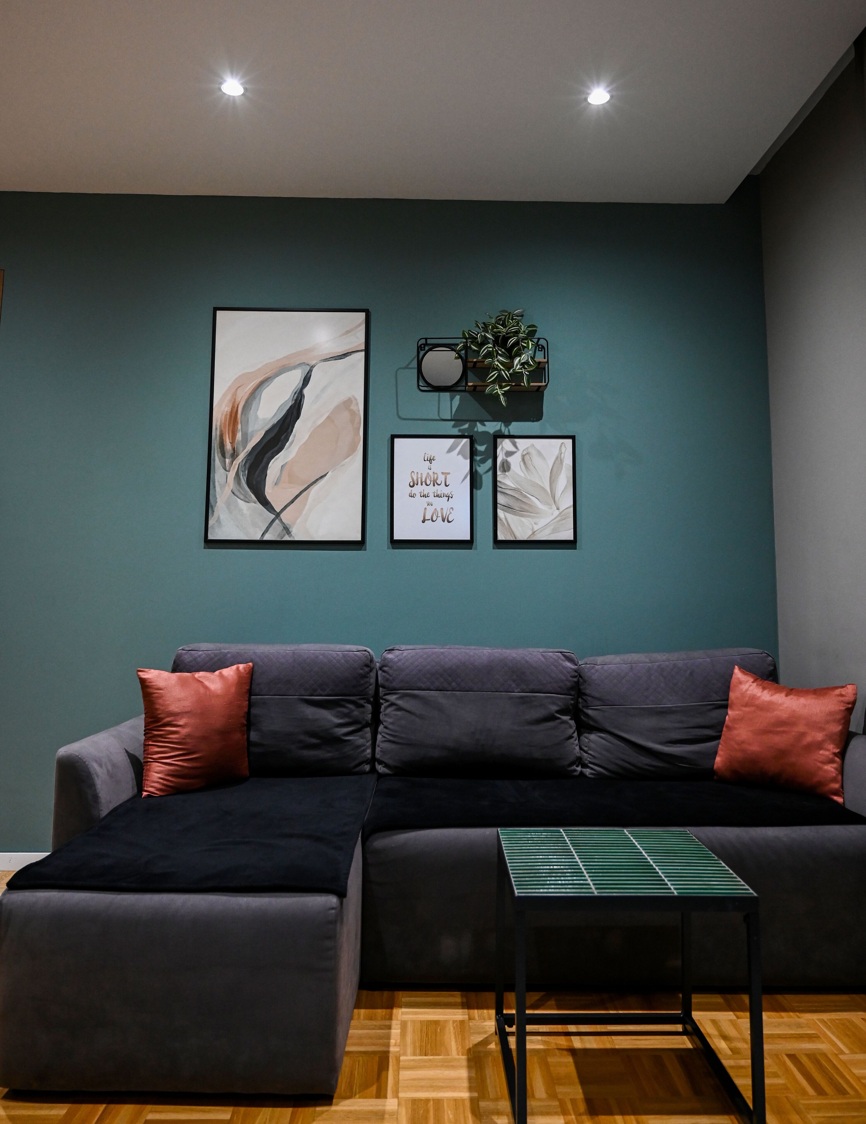 A modern living room with a dark grey sectional sofa, two red cushions, a small side table, and three framed artworks on the wall.