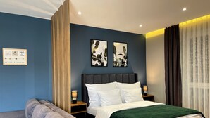 A modern bedroom with a dark blue wall, a large bed with white bedding, two bedside tables with lamps, a grey sofa, and a large window with sheer curtains. - Premium Apartments (Gjakova)
