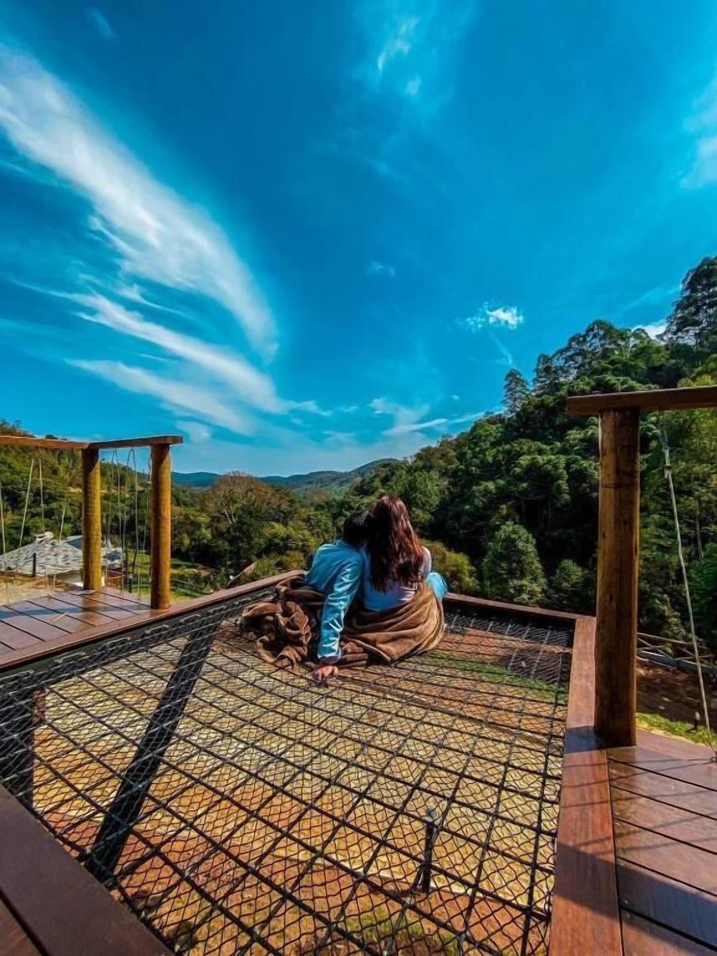 Una pareja sentada en una terraza de madera con una barandilla de seguridad de red, con vistas a un paisaje montañoso.