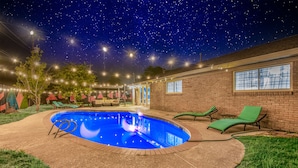 Un patio trasero con piscina, sillas reclinables verdes y luces colgantes.