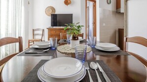 A dining area with a round table set for four, a television, a potted plant, and a kitchen visible in the background. - Gaia Verde by Interhome (San Vito lo Capo)