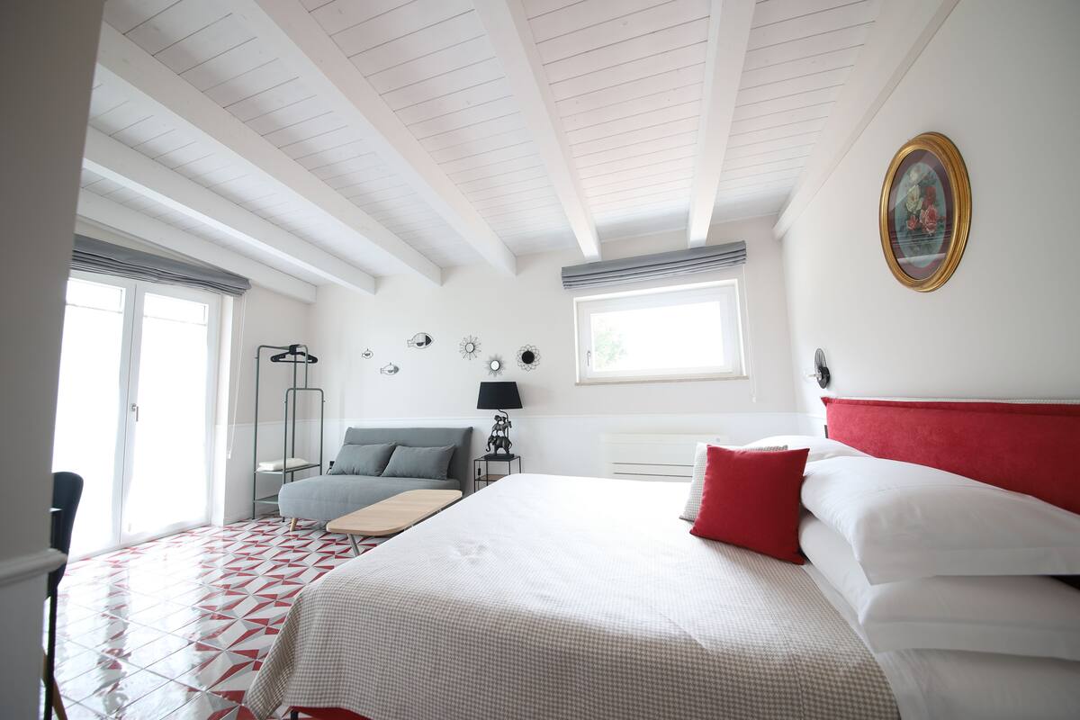 A hotel room with a large bed, a grey sofa, a small table, and a window with blinds.