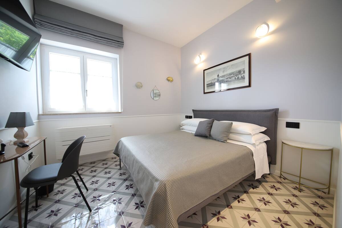 A bedroom with a bed, a desk, a chair, a window, and a wall-mounted picture.