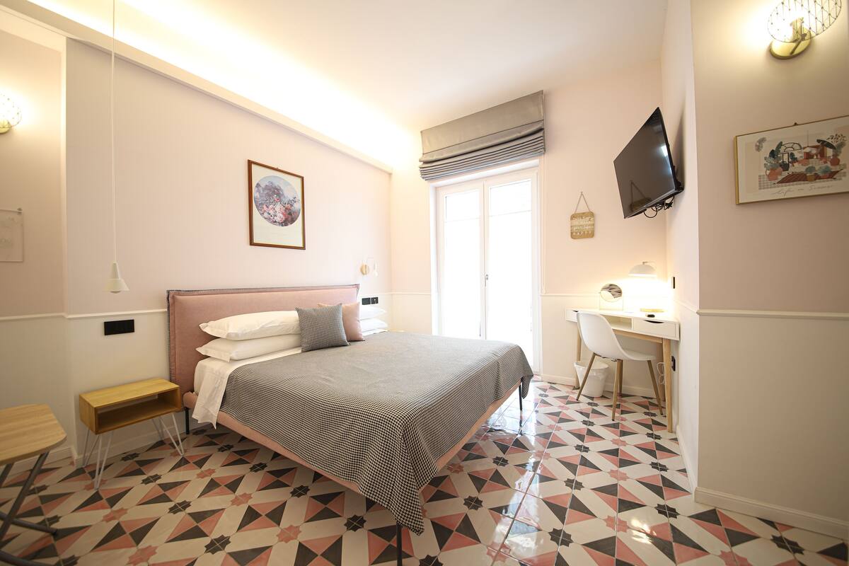 A bedroom with a bed, bedside table, desk, chair, and a TV mounted on the wall.