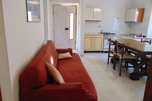 A living room with a red sofa, wooden dining table, and chairs, a kitchen with white cabinets and a stove, and a door leading to another room.