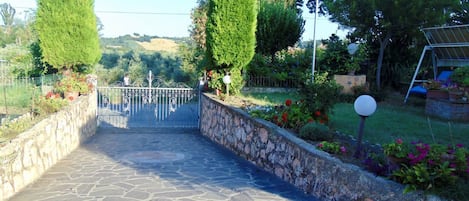 Un camí de pedra que condueix a una entrada tancada amb reixa, envoltada d'arbres i plantes amb flors ben podades.