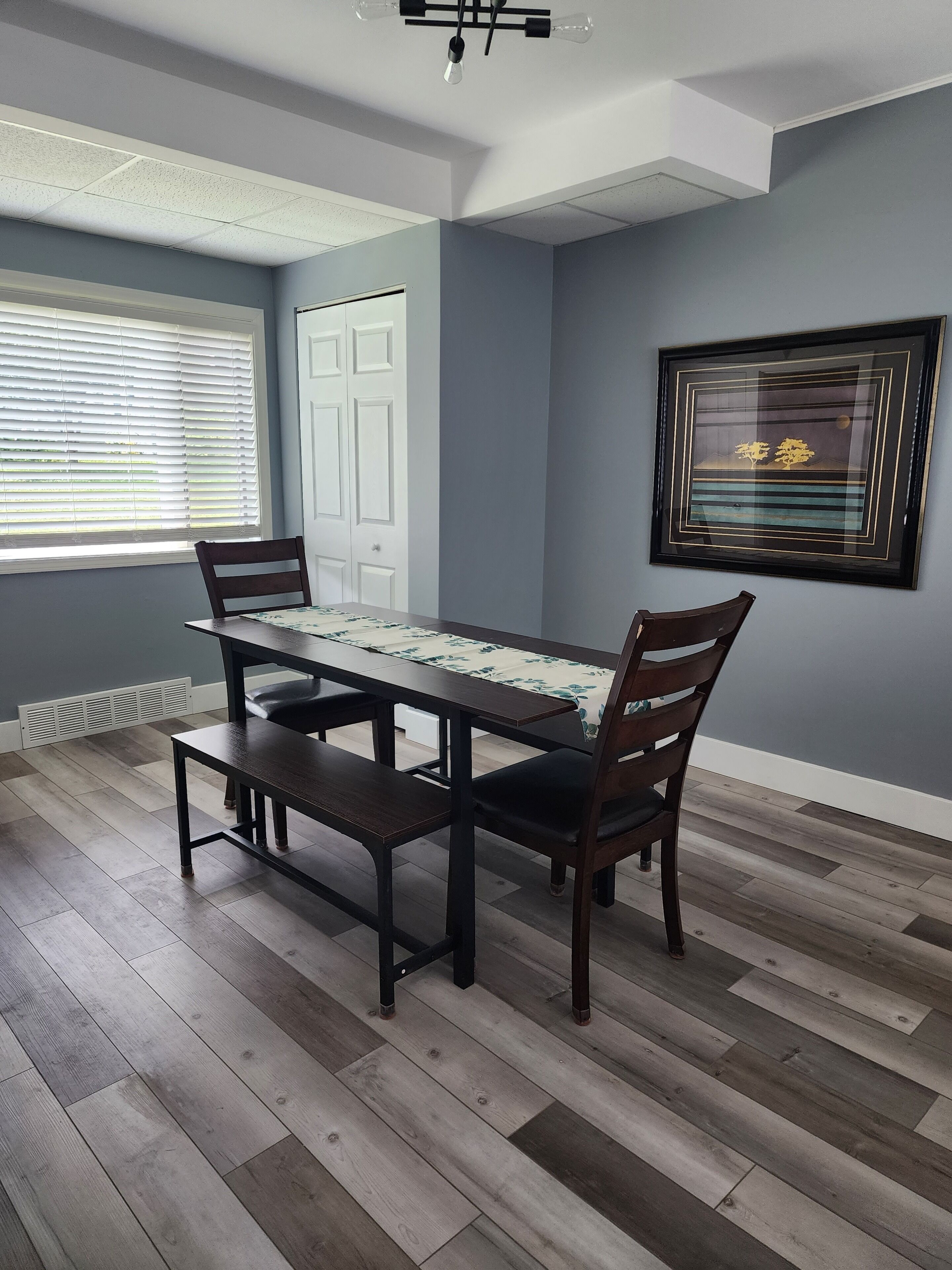 Un comedor con una mesa y sillas de madera oscura, un cuadro enmarcado en la pared y una ventana con persianas.