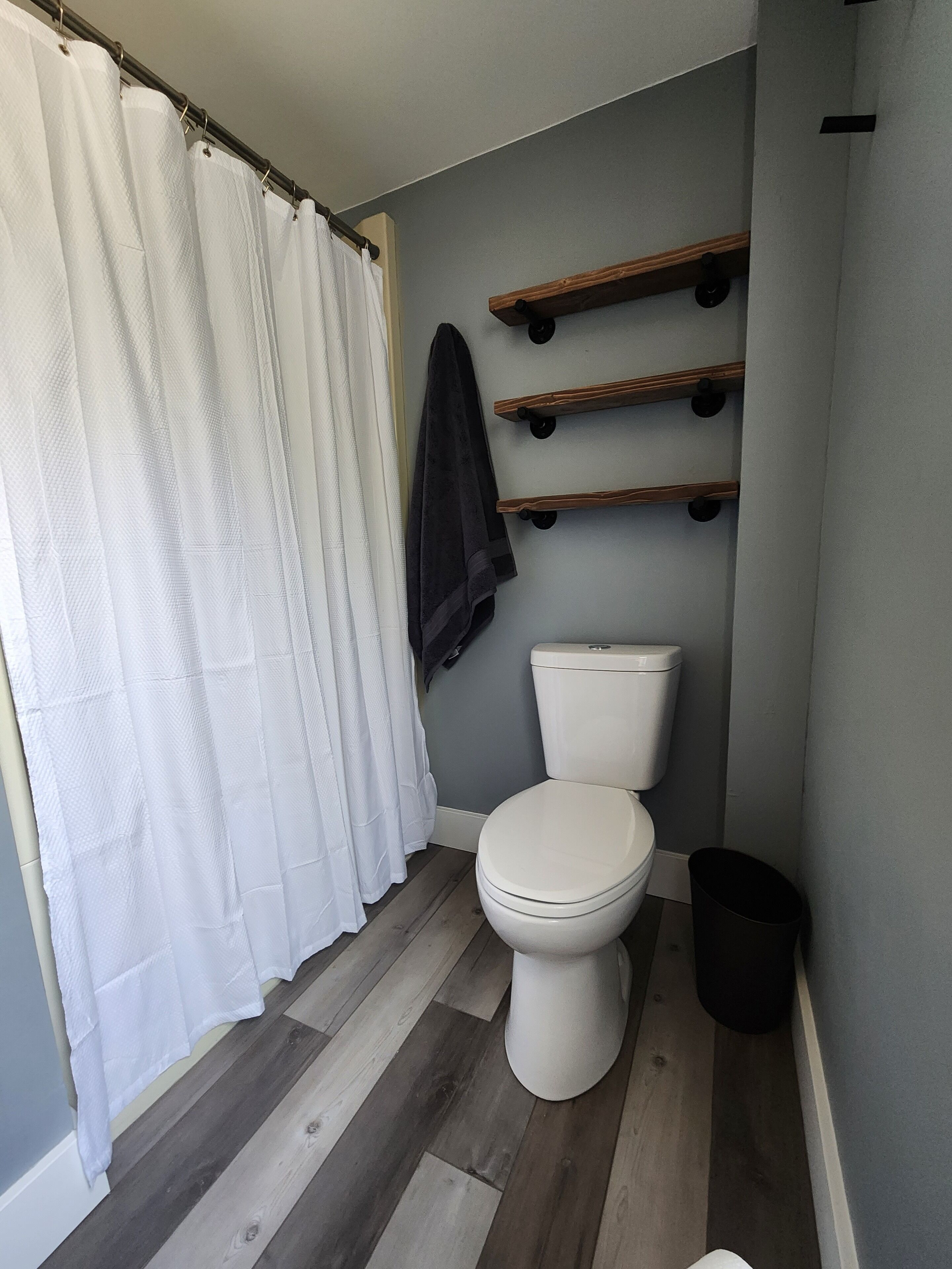 Een badkamer met een wit toilet, een douchegordijn, houten planken en een prullenbak.