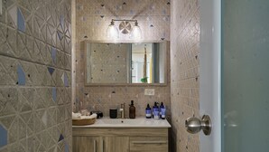 Une salle de bain avec un mur recouvert de carreaux aux motifs géométriques, un miroir au-dessus d’un lavabo agrémenté d’articles de toilette, et un meuble en bois.