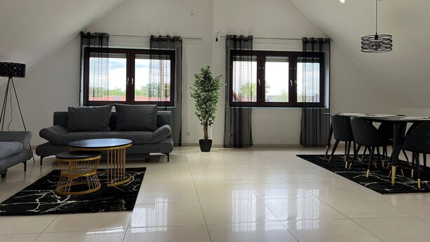 A spacious living area with a high ceiling, a grey sofa, a round coffee table, a dining table with chairs, and large windows with curtains.