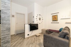 Uma sala de estar com televisão de tela plana na parede, um sofá cinza, piso de madeira e uma porta.