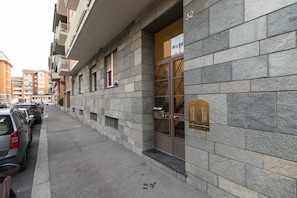 A multi-story building with a stone facade, a ground-floor entrance with a mailbox, and parked cars along the street.