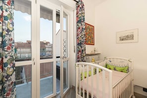 A room with a crib, a window with a view of buildings, a radiator, and a framed picture on the wall.