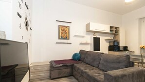 Ein modernes Wohnzimmer mit einem Ecksofa, einem Flachbildfernseher, einer Wanduhr und einer Küchenzeile im Hintergrund.