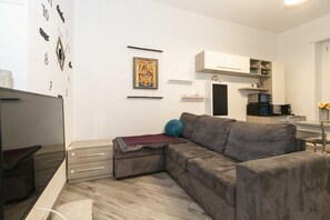 Ein modernes Wohnzimmer mit einem Ecksofa, einem Flachbildfernseher, einer Wanduhr und einer Küchenzeile im Hintergrund.