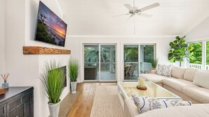 Amplia sala de estar con un sofá grande, una mesa de centro y un televisor montado en la pared. Cuenta con un ventilador de techo, un ventanal corredero que da a un patio exterior y varias plantas en macetas.