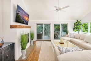 A spacious living room with a large sofa, a coffee table, and a television mounted on the wall. There is a ceiling fan, a sliding glass door leading to an outdoor patio, and several potted plants.