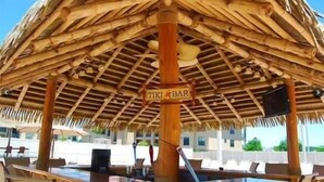 Eine strohgedeckte Struktur mit dem Schriftzug "TIKI BAR", Sitzgelegenheiten im Freien und einem Poolbereich.