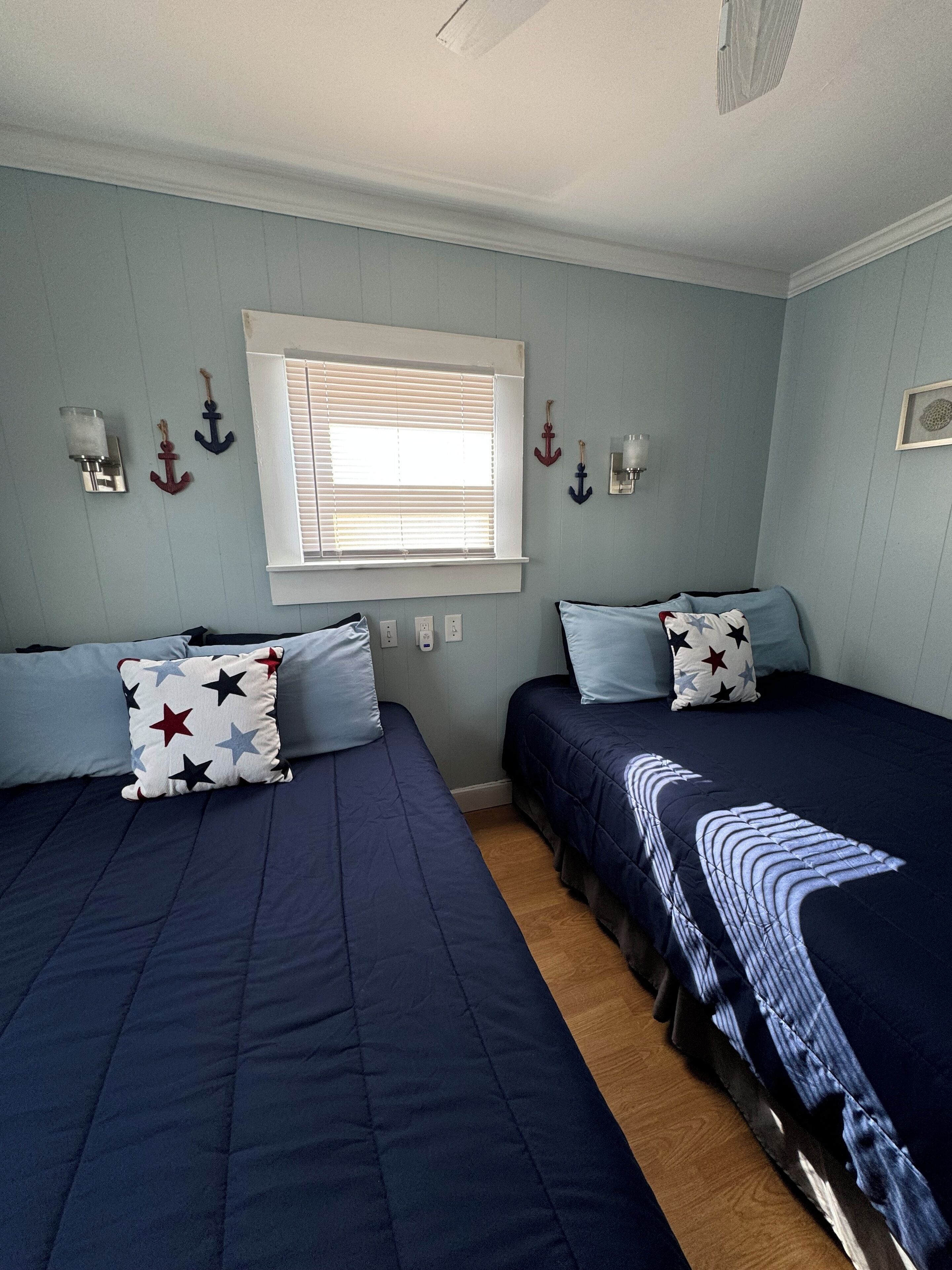 Habitación con dos camas, cada una con cubrecama y almohadas azules, una ventana con persianas, luces fijadas a la pared y un pequeño marco de fotos en la pared.