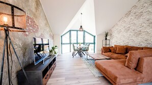 A modern living room with a high ceiling, exposed brick walls, a wooden floor, a brown sofa, a dining table with chairs, and a TV on a stand. - Relax in our (Losheim am See)