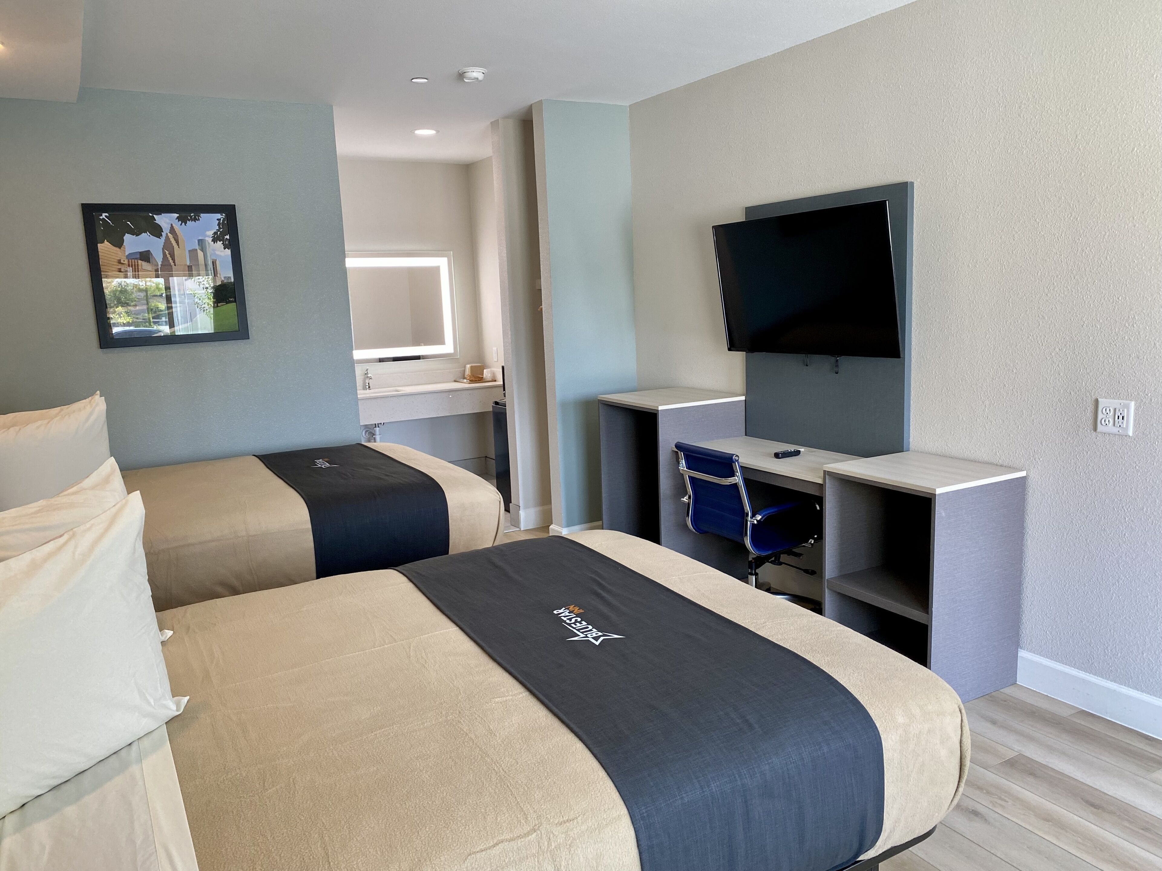 Habitación de hotel con dos camas, un escritorio, una silla, un televisor y un baño visible al fondo.