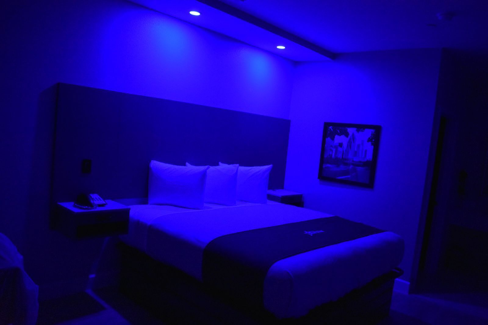 Una habitación de hotel con una cama, una mesita de noche, un cuadro en la pared y una lámpara en el techo.