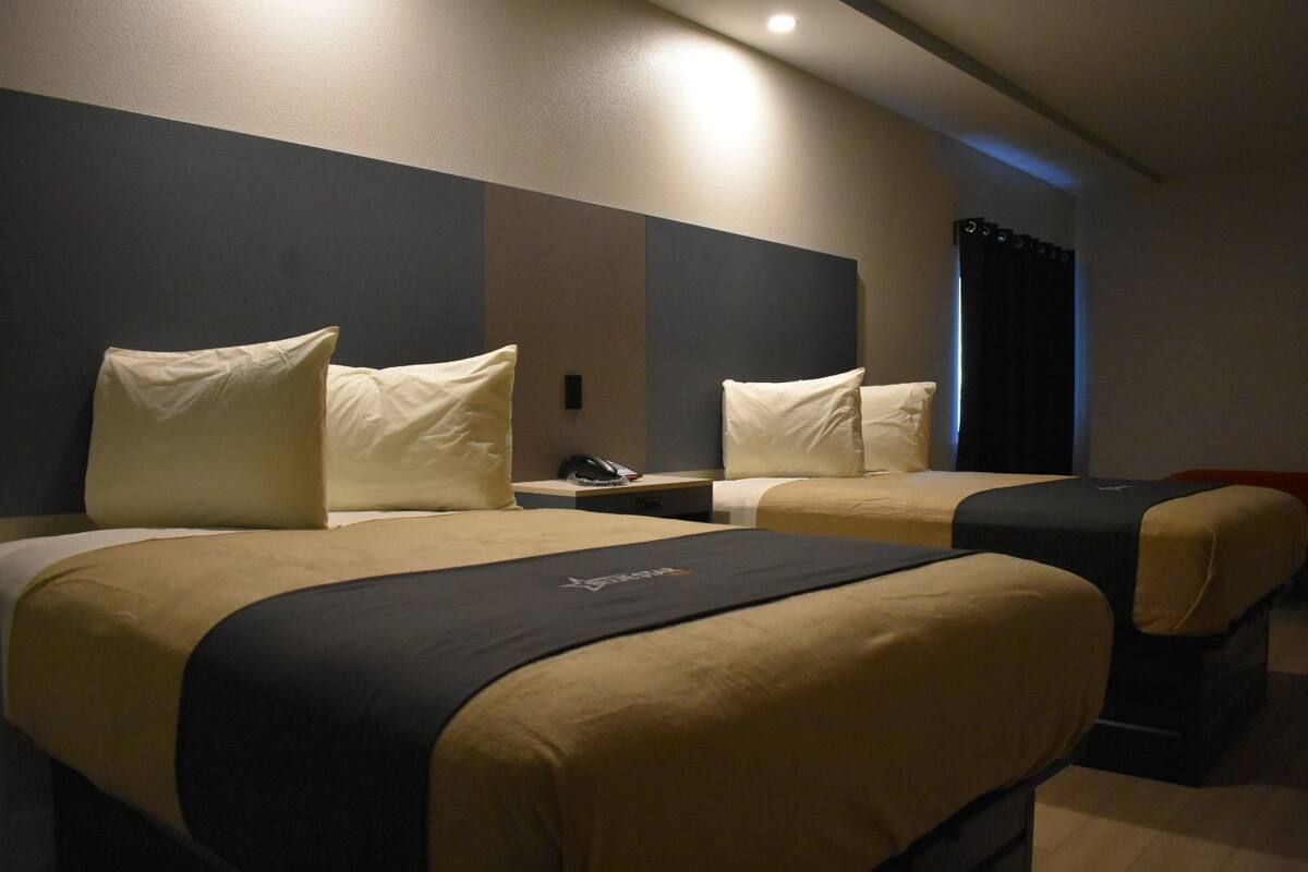 A hotel room with two beds, each with a black headboard, beige bedding, and a small bedside table with a lamp.
