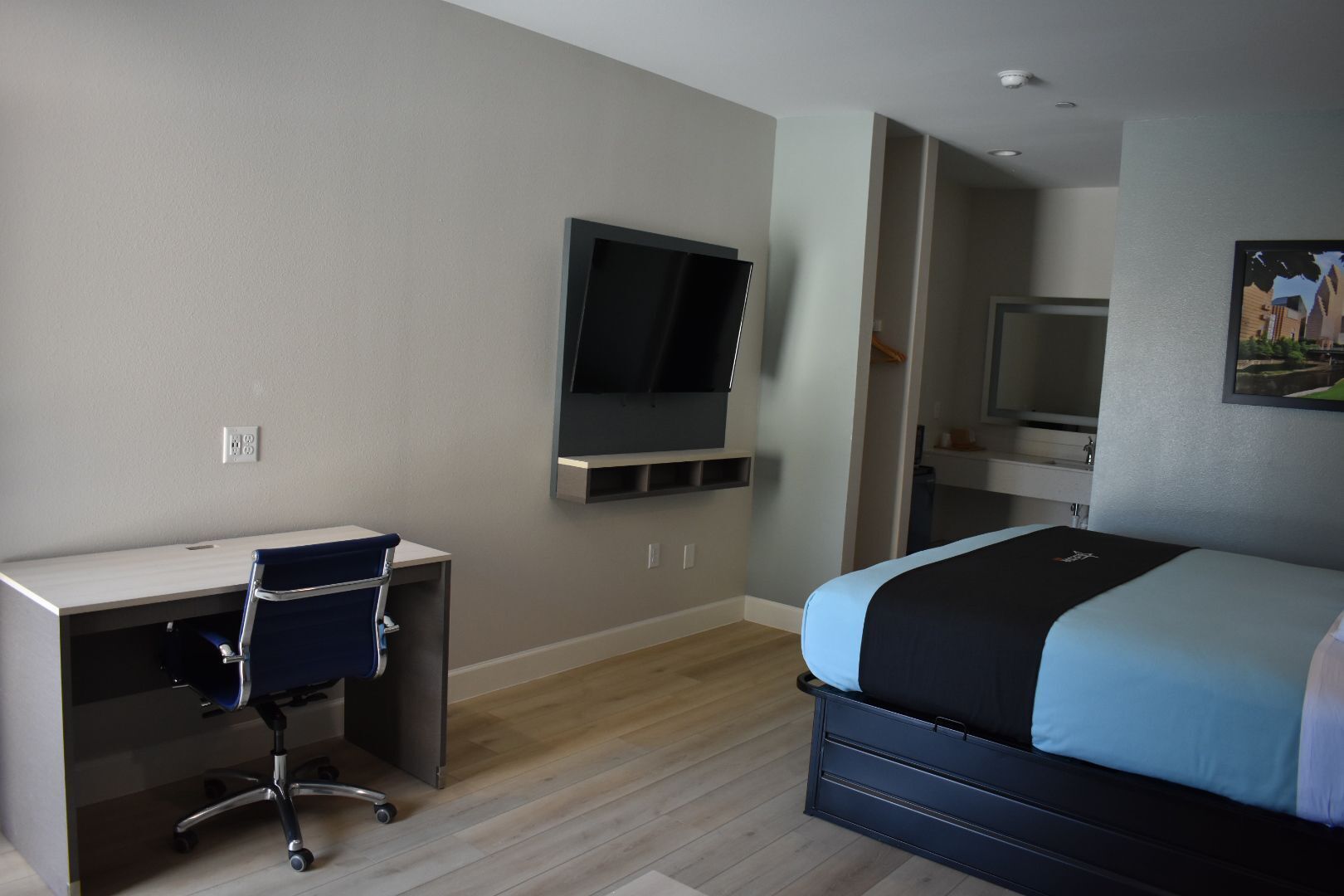 A modern hotel room with a desk, chair, bed, TV, and wardrobe.