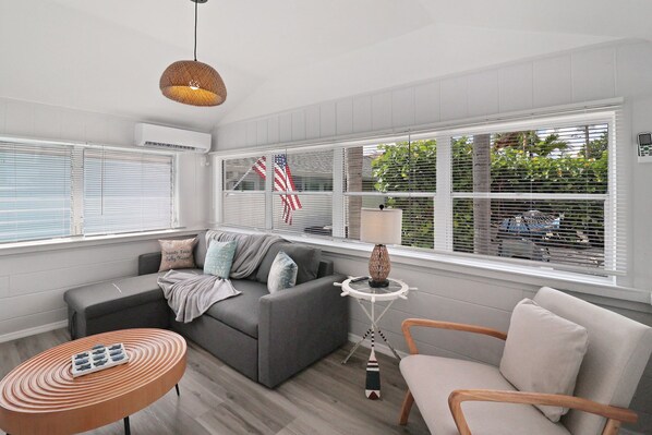A modern living room with a grey sofa, a round wooden coffee table, and a wooden chair. There is a hanging lamp, a small side table with a lamp, and a window with blinds.