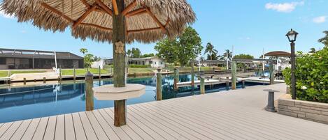 A wooden deck with a thatched umbrella, a pool, and a view of buildings in the background.