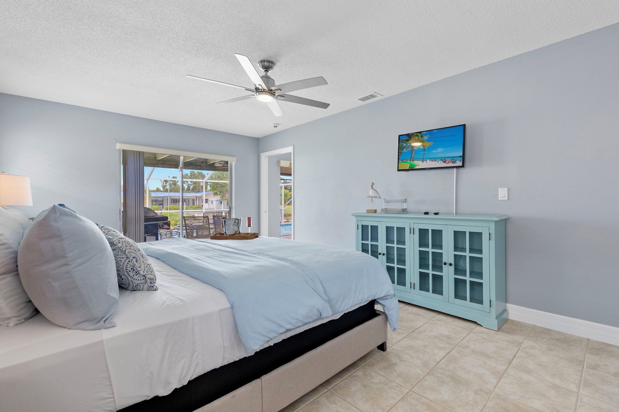 A bedroom with a bed, a ceiling fan, a TV, a cabinet, and a view of the outside.