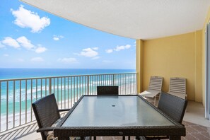 Un balcon avec une table en verre et des chaises en osier, donnant sur une plage et l’océan.