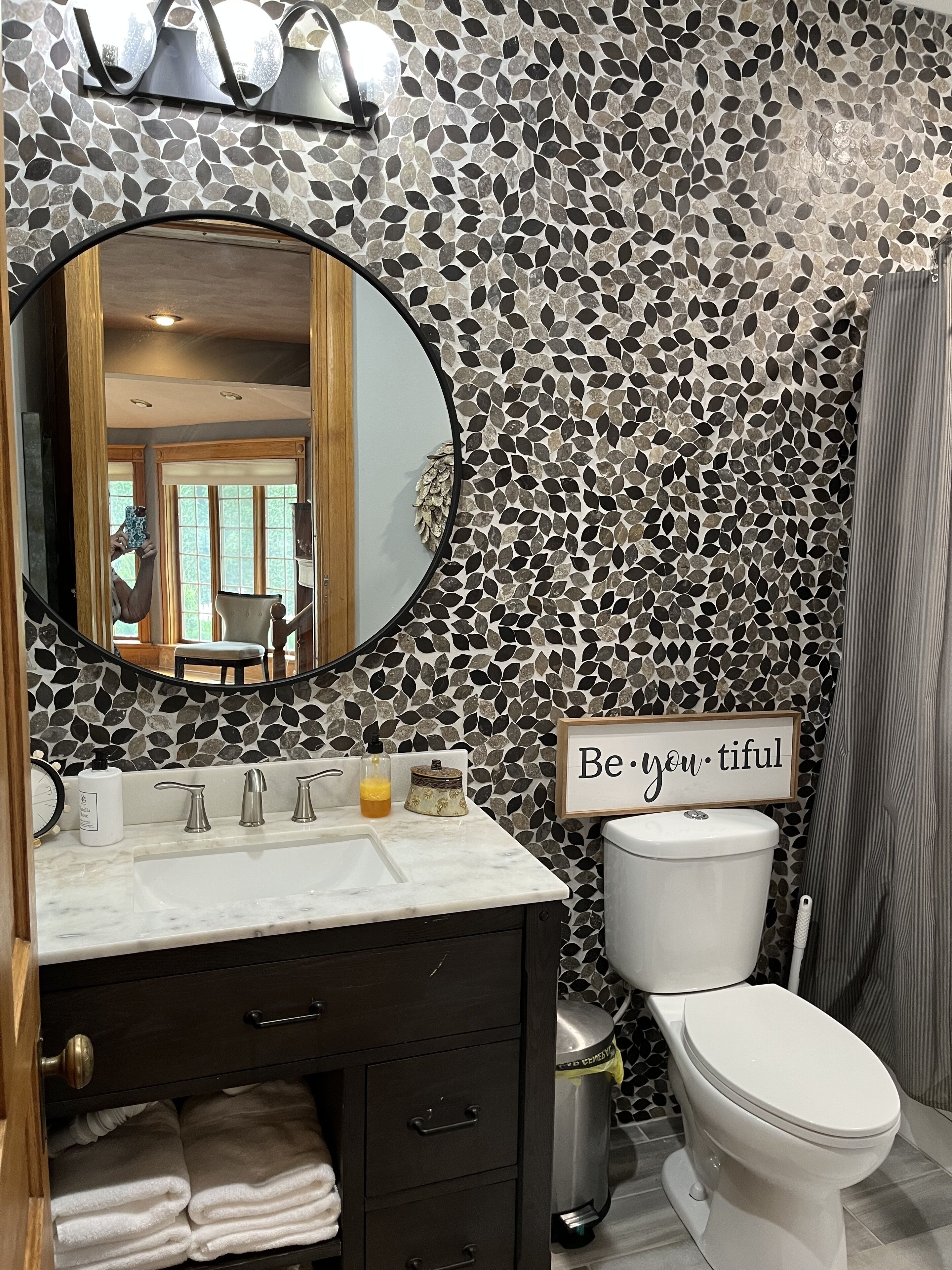 A bathroom with a mosaic tile wall, a round mirror, a sink with a marble countertop, a toilet, and a sign that says "Be you, tiful".