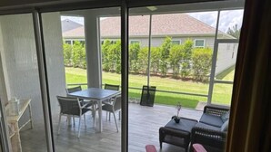 Un porche techado con mesa y sillas, un rincón de estar con sofá y sillones, y vistas a un césped verde y casas al fondo.