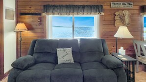 A cozy living room with a view of a lake, a sign indicating the location, and a couch with a 'camp' pillow. - Kineo Point (Waterfront on Moosehead Lake) (Rockwood)