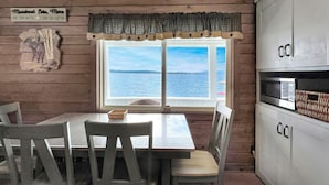 A dining area with a table and chairs, a window with a view of the water, and a kitchen area with cabinets and a microwave. - Kineo Point (Waterfront on Moosehead Lake) (Rockwood)