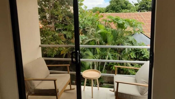 A balcony with two rocking chairs and a small table, overlooking a lush garden and a red-roofed building.