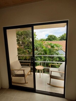 Un balcón con dos mecedoras y una pequeña mesa, con vistas a un jardín exuberante y un edificio de techo rojo.
