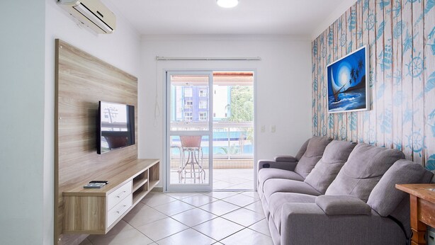 A modern living room with a flat-screen TV, a grey sofa, a wooden TV stand, and a sliding glass door leading to a balcony.