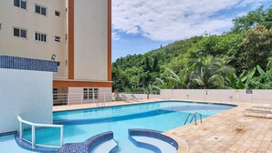 A swimming pool with a tiled deck, surrounded by a building with a tiled exterior and a lush green hillside in the background.