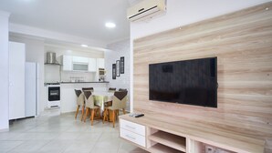 A modern kitchen with white cabinets, a microwave, and an oven. There is a dining area with a table and chairs. A flat-screen TV is mounted on a wooden wall.