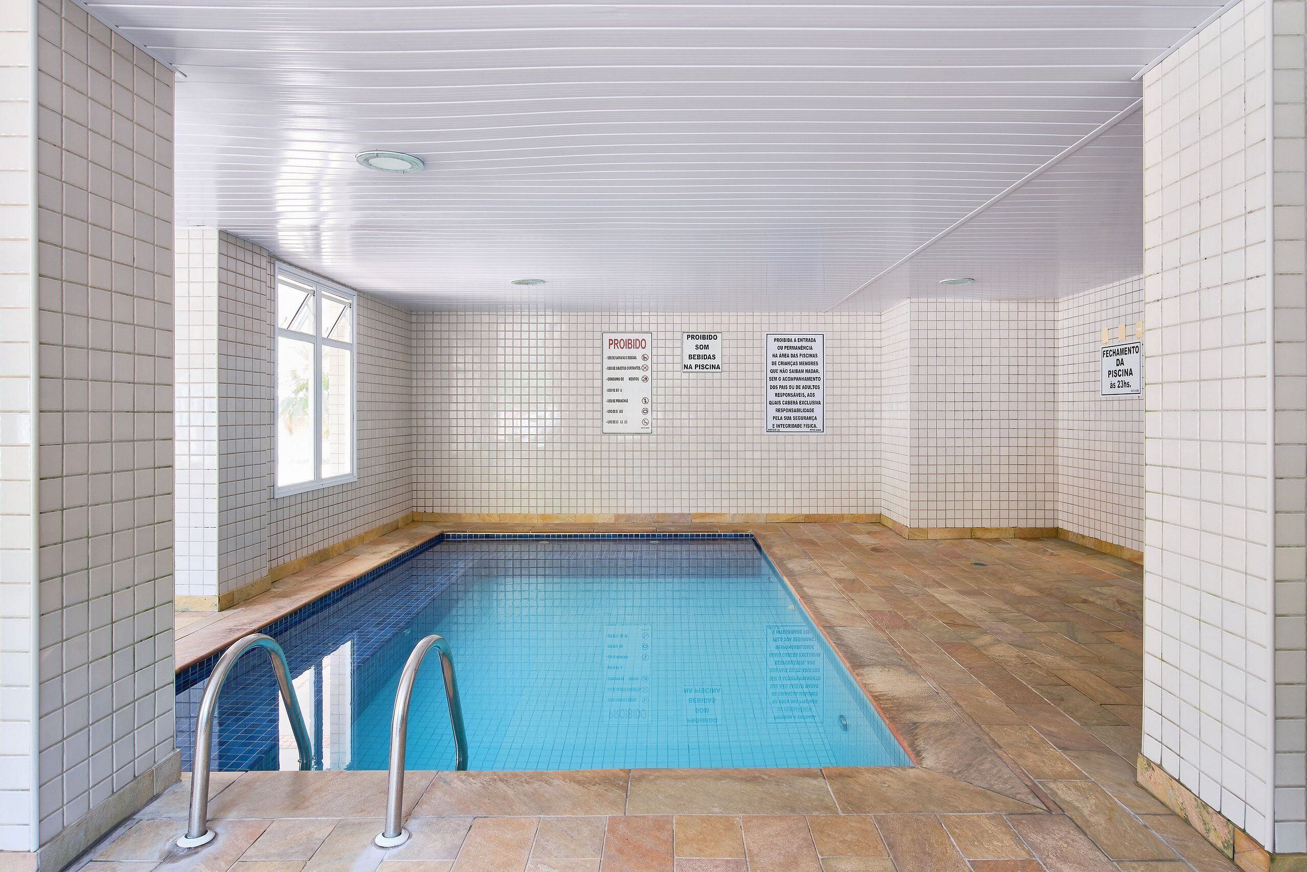 Une piscine intérieure avec des murs et un sol carrelés, des marches d’accès à la piscine bien visibles et plusieurs panneaux d’information.