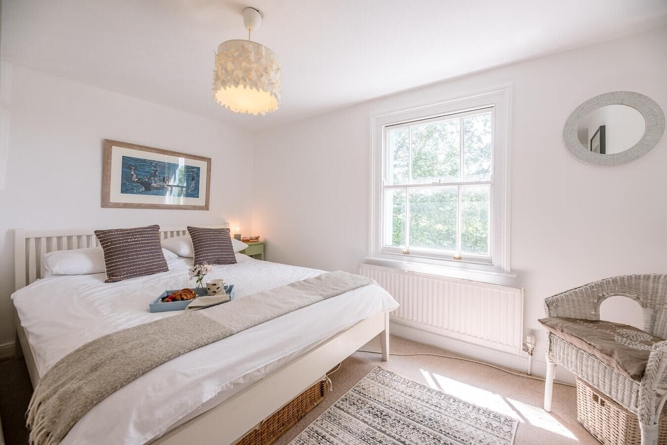 A bedroom with a large bed, a bedside table, a wicker chair, a window with a view of greenery, and a ceiling light fixture.