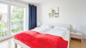 A bedroom with a large bed, white bedding, a red throw blanket, a nightstand with a lamp, and a view of greenery through a sliding glass door. - Traum-ferienwohnung Alpenblick, 75 qm, Balkon und Bergblick (Bad Aibling)