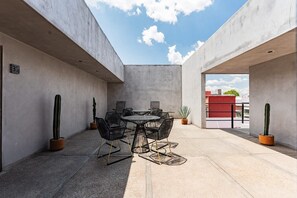 Eine Dachterrasse mit rundem Tisch und Stühlen, umgeben von Kübelkaktussen, unter blauem Himmel.